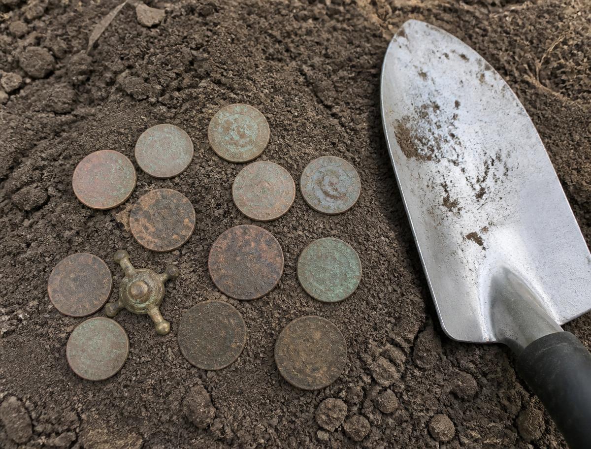 Dug copper coins and a small brass relic laid out on soil next to a hand trowel